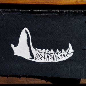 May include: A rectangular black fabric patch with frayed edges featuring a white screen-printed graphic of a jawbone with sharp teeth. Below the teeth, the text reads Bᛟᛚᛗ:ᚺᚾᛏ:ᛗᚱ in a runic-style font. The patch is secured with pins at the top corners.