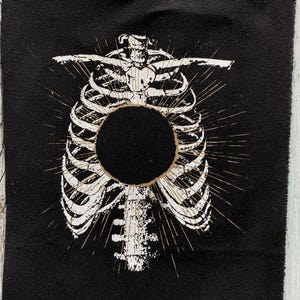 May include: Black fabric patch with a white skeletal rib cage design and a central black circle. The rib cage has a detailed, aged appearance, with radiating lines from the centre. The edges of the patch are unfinished.