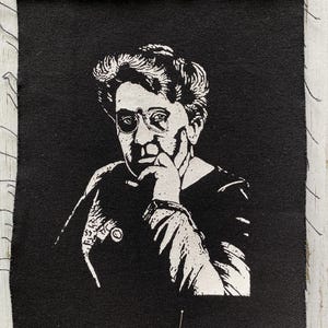 May include: Black fabric patch featuring a white portrait of a person with glasses, resting their chin on their hand. The portrait is a detailed illustration. The edges of the patch are frayed. A pin is inserted into the fabric.