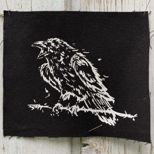 Op de afbeelding: Zwart vierkantige patch met een witte illustratie van een raaf die op een tak zit. De raaf wordt afgebeeld met zijn snavel open, alsof hij kraait. De patch heeft een rauwe rand, waardoor hij een handgemaakt uiterlijk heeft. De raaf is een symbool van mysterie en intelligentie.