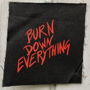 May include: A square black fabric patch with raw, frayed edges. The text BURN DOWN EVERYTHING is printed in a bold, bright red, hand-drawn style font across the center of the patch. The fabric has a visible woven texture throughout the surface.