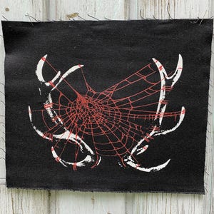 May include: Black patch with a white and red spiderweb design. The web is centered with a detailed pattern, and the white design resembles antlers or horns. The patch has a raw edge.