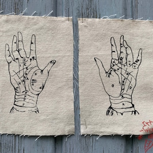 May include: Two rectangular pieces of beige canvas fabric, each featuring a black ink illustration of a human palm with palmistry lines and various letters printed on the skin. The edges of the fabric are frayed, and the text Art Light Werkstatt is visible.
