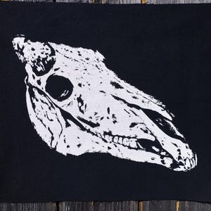 Könnte beinhalten: Ein quadratischer schwarzer Stoffaufnäher mit einer kontrastreichen, weiß siebgedruckten Grafik eines Pferdeschädels. Der Schädel ist im Profil dargestellt und zeigt die Augenhöhle, Nasenhöhle und Zähne mit einer texturierten Optik.
