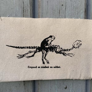Pode incluir: Um patch retangular de tecido bege com uma ilustração preta de um esqueleto de rã montando um lagarto esquelético. O texto "Crapaud se rendant au sabbat" está impresso abaixo. O patch está preso a uma superfície de madeira desgastada.