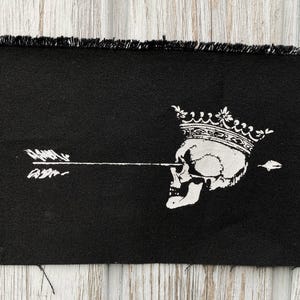 May include: Black fabric patch with a white graphic of a skull wearing a crown, pierced by an arrow. The edges of the patch are frayed. The skull and arrow design is centered on the patch.