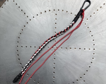 May include: A lanyard featuring a braided cord in black, white, and red, finished with black heat-shrink tubing at both ends. A black metal carabiner is attached to one end, and two loose red cords extend from the same attachment point.