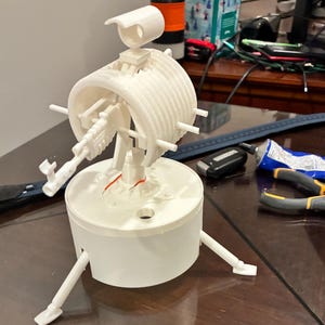 May include: A white 3D-printed model of a futuristic device. The model features a cylindrical core with a gun-like structure, supported by three legs. A smaller, rectangular component is mounted above the cylinder.