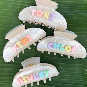 May include: Four white pearlescent hair claw clips featuring pastel-colored letter beads. The clips are customized with the names TORI, KIKI, CHLOE, and ASHLEY in raised, multi-colored lettering. Each clip has a spring-loaded mechanism and teeth.