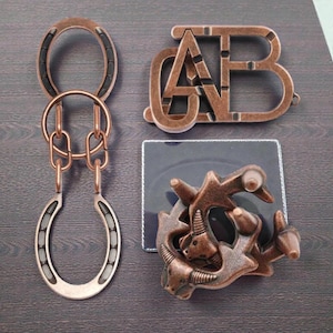 Puede incluir: Tres rompecabezas de metal de color cobre. Un rompecabezas presenta dos herraduras conectadas por cadenas. Otro rompecabezas tiene la forma de las letras "CAB". El tercer rompecabezas tiene un diseño complejo con figuras de animales.