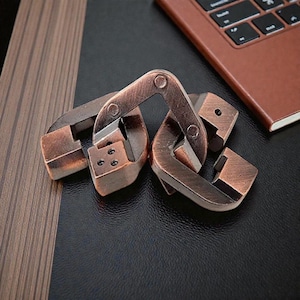 Pode incluir: Um quebra-cabeça de metal cor de bronze com peças interligadas. O quebra-cabeça é feito de vários componentes metálicos retangulares e curvos. O quebra-cabeça está em uma superfície preta, com um laptop marrom ao fundo. É um desafio mental.