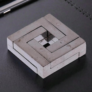 May include: A silver metal puzzle with a square shape. The puzzle is composed of several interlocking pieces, creating a complex design. The center of the puzzle has a smaller square opening. The puzzle is on a dark surface.