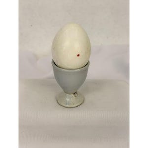 May include: A smooth, off-white egg with a single small red speck sits upright in a light gray, pedestal-style ceramic egg cup. The egg cup features a rounded bowl shape supported by a short, flared base, providing a stable stand for the egg.