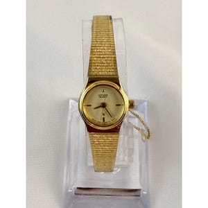 May include: A gold-toned Citizen Quartz watch with a rectangular gold band. The watch face is gold with black hands and markers. The watch is displayed on a clear plastic stand.