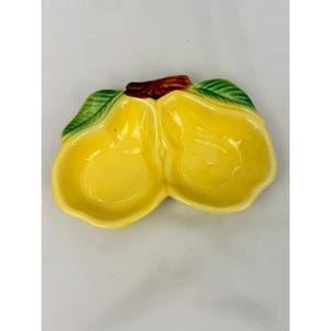 May include: A ceramic dish shaped like two conjoined yellow pears with a brown stem and green leaves at the top. The interior of the dish is smooth and yellow, while the exterior features a glossy finish with defined pear-shaped indentations.