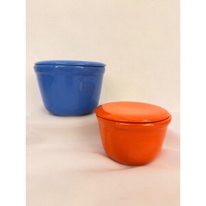 May include: Two ceramic bowls with matching lids, one in a vibrant blue and the other in a bright orange. Both containers feature a smooth, glossy finish, a rounded base, and a simple, clean design with a fitted lid resting on top.