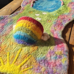 Könnte beinhalten: Ein farbenfrohes Filz-Spielplatz-Matte mit Regenbogen, Sonne und blauem See. Die Matte hat eine grüne Basis mit gestickten Details von Blumen und Wolken. Der Regenbogen besteht aus rotem, orangefarbenem, gelbem, grünem, blauem und violettem Filz.
