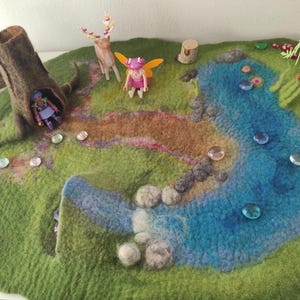 Può includere: Un tappetino da gioco in feltro raffigurante una scena fatata stravagante con un lago blu, erba verde e un tronco d'albero marrone. Miniature di una fata, un cervo e una persona sono posizionate sul tappetino, insieme a pietre decorative di vetro.