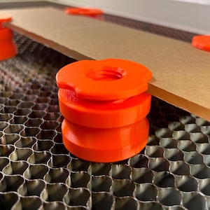 May include: A set of bright orange, circular, 3D-printed honeycomb bed pins with a scalloped edge design. These spacers feature a central hole and a grooved side profile, positioned to support a flat, rectangular brown material sheet on a metal grid.