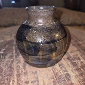 May include: A small, handcrafted ceramic vase with a rounded body and a narrow neck. The vase features a dark brown exterior with streaks of blue and yellow. The pottery has a textured, speckled appearance.