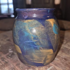 Peut inclure: Un vase en céramique fait à la main, à large ouverture et corps arrondi. Le vase présente un mélange de couleurs bleu, jaune et turquoise, avec un bord bleu foncé. L'extérieur a un aspect texturé et peint, avec un petit symbole infini gravé sur le côté.