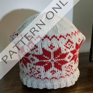 Può includere: Un cappello di lana grigio e rosso con un motivo a fiocco di neve. Il cappello è realizzato con un filato morbido e ha un bordo a coste.