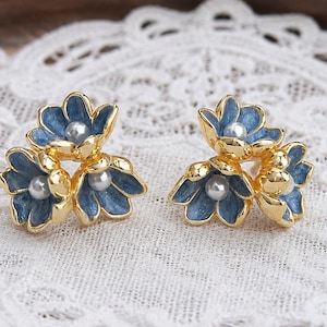 Può includere: Un paio di orecchini a lobo dorati, ciascuno composto da tre piccoli fiori con petali in smalto blu e una perla bianca centrale. Il design floreale è disposto a grappolo, con una finitura metallica lucida sui bordi dei petali.
