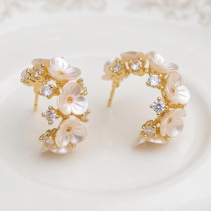 Peut inclure: Une paire de boucles d'oreilles créoles dorées ornées de petites fleurs blanches et de pierres transparentes scintillantes. Les boucles d'oreilles présentent une forme semi-circulaire avec un fermoir à tige, formant un arrangement floral.