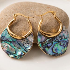 May include: A pair of gold hoop earrings featuring a semi-circular inlay of iridescent abalone shell. The earrings have a polished gold metal frame with a hinged closure mechanism, showcasing a vibrant mix of blue, green, and purple shell patterns.