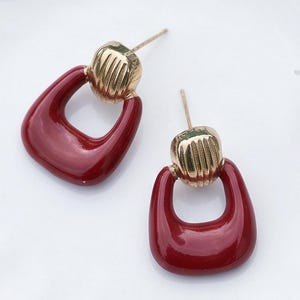 May include: A pair of stud earrings featuring a gold-toned metal top with vertical ridges and a smooth, deep red, U-shaped dangling component. The earrings are displayed on a white surface with a subtle floral pattern and white flower accents.