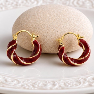 May include: A pair of small hoop earrings featuring a twisted design with a deep red enamel finish and gold-toned metal edges. The earrings have a hinged closure mechanism and a polished, reflective surface that catches the light.