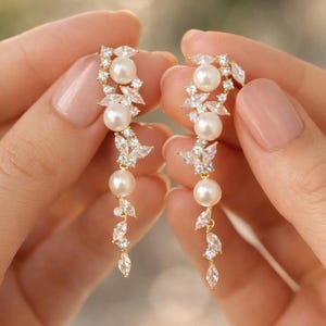 Puede incluir: Un par de pendientes dorados con tres perlas blancas y cristales transparentes. Los pendientes tienen un diseño floral con elementos colgantes, ideales para ocasiones especiales.