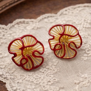 May include: A pair of circular stud earrings featuring a gold-tone wire mesh interior framed by a bold, glossy red enamel outline. The design resembles a stylized flower or abstract organic shape with radiating lines extending from the center point.