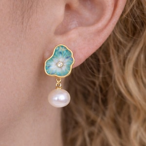 May include: Gold earrings with a floral design, turquoise and white enamel, and a small diamond. Each earring has a dangling white pearl. The earrings are displayed on a person's ear.