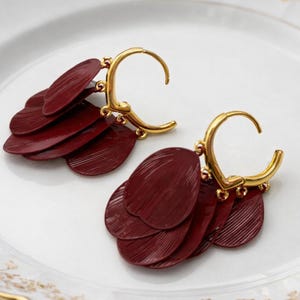 May include: A pair of gold-tone hoop earrings featuring multiple dangling, oval-shaped, deep burgundy red charms with a textured, ribbed surface. The charms are attached to the base of each hoop, creating a layered, fan-like appearance.