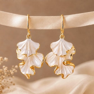 Pode incluir: Um par de brincos pendentes dourados com pingentes brancos em forma de pétalas sobrepostas, com superfícies lisas e brilhantes e bordas douradas finas. Os brincos pendem de argolas douradas simples, criando um visual floral delicado.