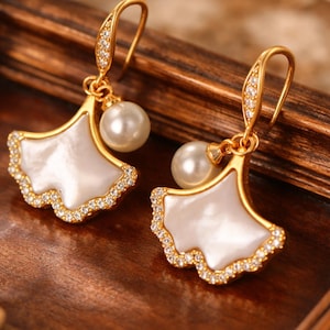 Peut inclure: Une paire de boucles d'oreilles pendantes dorées présentant un motif de feuille de ginkgo en éventail avec une incrustation de nacre blanche, rehaussée de petits cristaux clairs sur le bord et d'une perle blanche ronde suspendue.