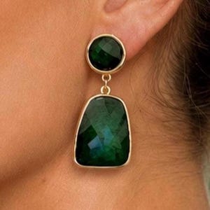 Puede incluir: Un pendiente colgante de tono dorado que presenta una pequeña piedra preciosa redonda facetada de color verde oscuro en la parte superior, unida por un fino eslabón metálico a una piedra facetada trapezoidal más grande.
