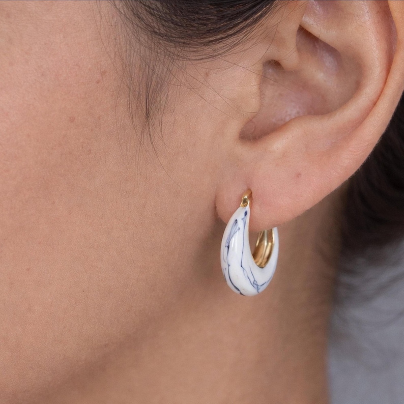 May include: A gold hoop earring with a white and blue marbled design. The earring is a teardrop shape and is worn on the ear. The earring is a stylish accessory.