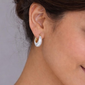 May include: A close-up of a person's ear wearing gold hoop earrings. The earrings feature a white and blue marbled pattern on the outer surface. The inner part of the hoop is gold.