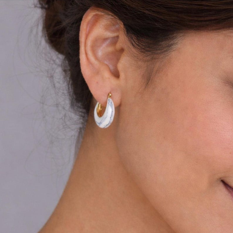 May include: A close-up of a person's ear wearing gold hoop earrings. The earrings feature a white and blue marbled pattern on the outer surface. The inner part of the hoop is gold.