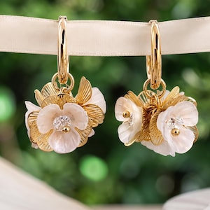 May include: A pair of gold hoop earrings featuring dangling floral charms. Each charm consists of layered white translucent petals, textured gold-tone metal leaves, and a small clear crystal center. The earrings hang from a smooth gold hoop.