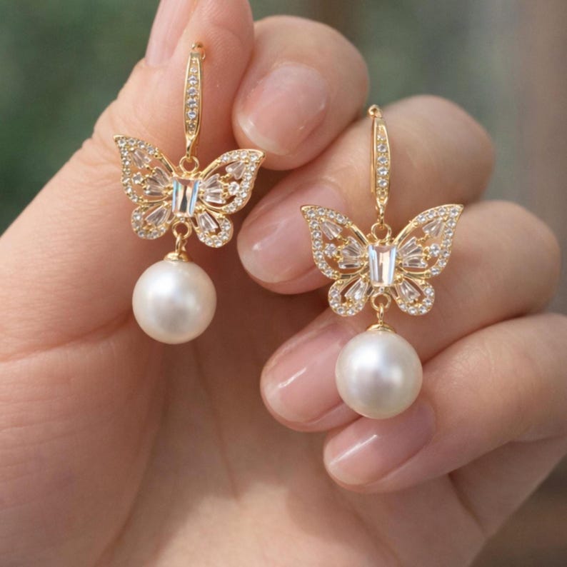 Pu&ograve; includere: Orecchini a farfalla color oro con cristalli trasparenti e una grande goccia di perla bianca. Le ali della farfalla sono decorate con piccoli cristalli. Gli orecchini sono tenuti da un gancio color oro.