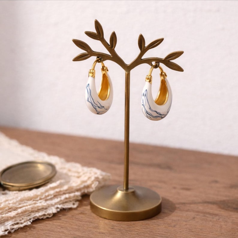 May include: White and blue marbled hoop earrings with gold detailing, presented on a gold-coloured metal earring stand. The earrings feature a distinctive, organic pattern and a polished finish. The stand is shaped like a tree branch.