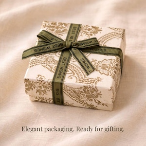May include: A small, square gift box with a white background and a gold paisley pattern. The box is tied with a green ribbon that reads "BEST WISHES" in white. The text "Elegant packaging. Ready for gifting." is at the bottom.