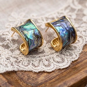 Peut inclure: Une paire de boucles d'oreilles créoles dorées avec une incrustation de nacre d'ormeau irisée. Les morceaux de coquillage forment un motif en mosaïque aux teintes changeantes de vert, bleu et violet. Les boucles ont une forme large et incurvée.