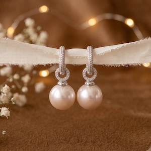 May include: A pair of drop earrings featuring large, round, cream-coloured pearls suspended from silver-toned hoops. Each hoop is encrusted with small, clear, sparkling stones that form a circular setting connecting the pearl to the earring post.
