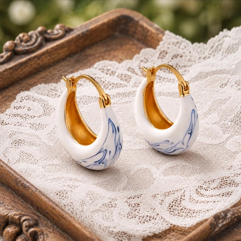 May include: A pair of gold-coloured hoop earrings with a white ceramic finish. The earrings feature a blue swirl design and a gold interior. The earrings are displayed on a white lace surface.
