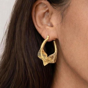 May include: Gold-toned hoop earrings with an intricate, open design. The earrings have a spiral and linear pattern, creating a textured appearance. The earrings are displayed on a person's ear, highlighting their shape and style.