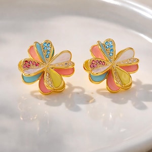 May include: A pair of gold-toned clover-shaped stud earrings. Each earring features five colorful petals in shades of pink, yellow, blue, and white, accented with small gemstones. The earrings have a gold-colored border and are set against a white background.